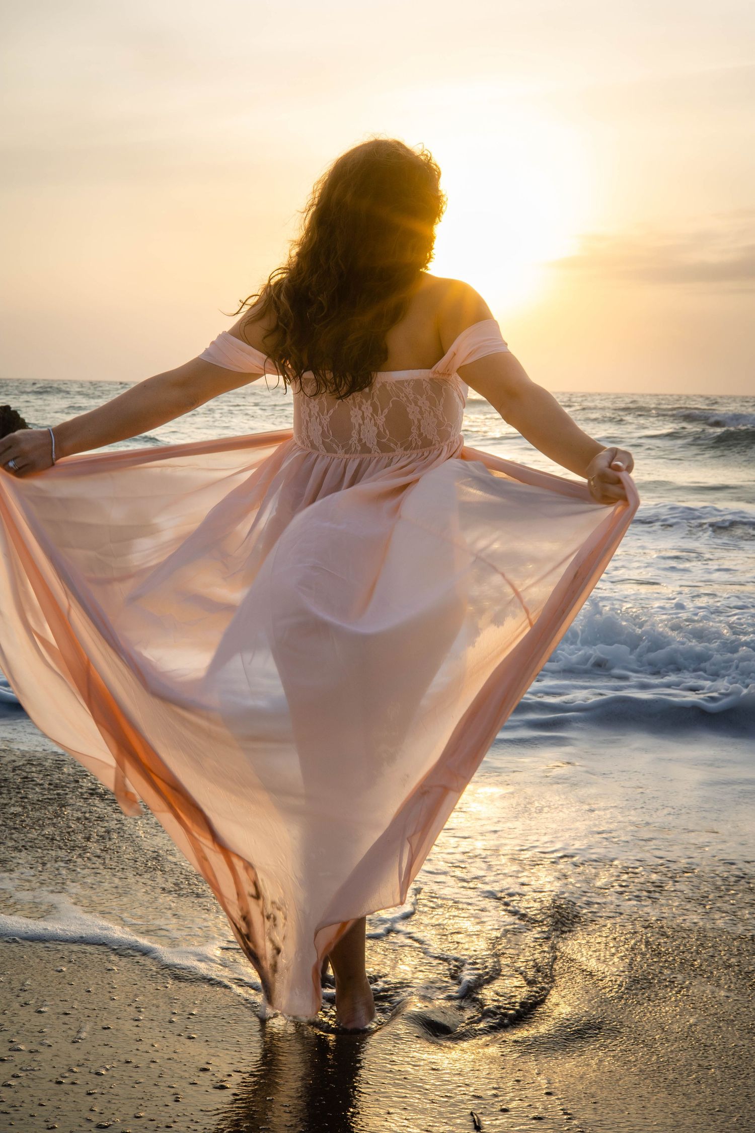 A woman in a sheer pink dress is facing a beautiful golden hour at sunrise.