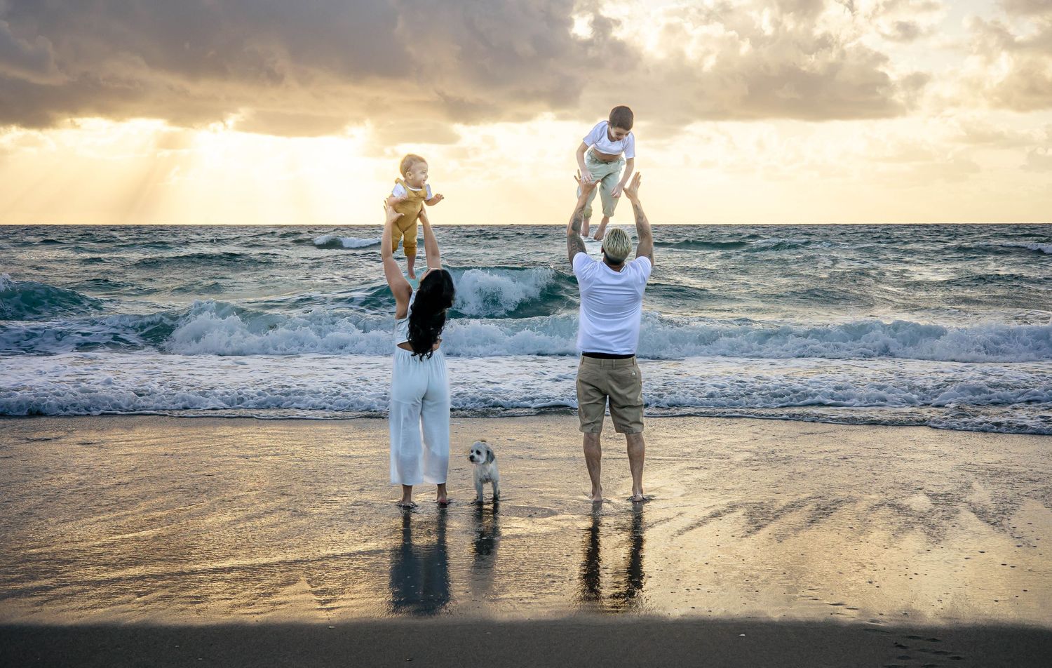 Family of four enjoying a beautiful sunset on the beach, parents lifting toddlers in the air by the ocean waves.