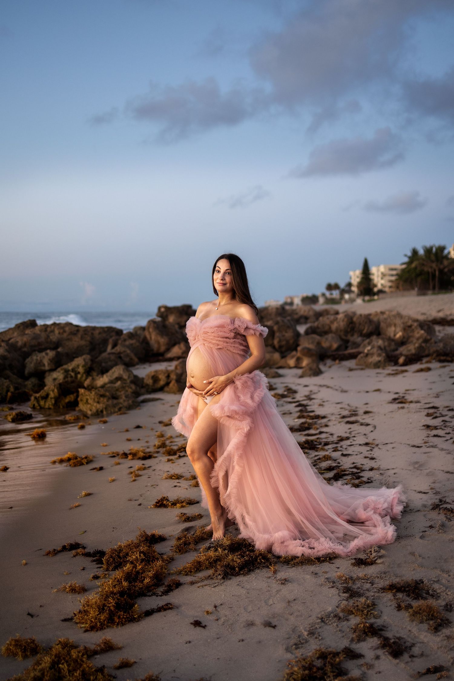 Pregnant woman in pink tulle gown poses on sandy beach at sunset with rocky shoreline and ocean in background.