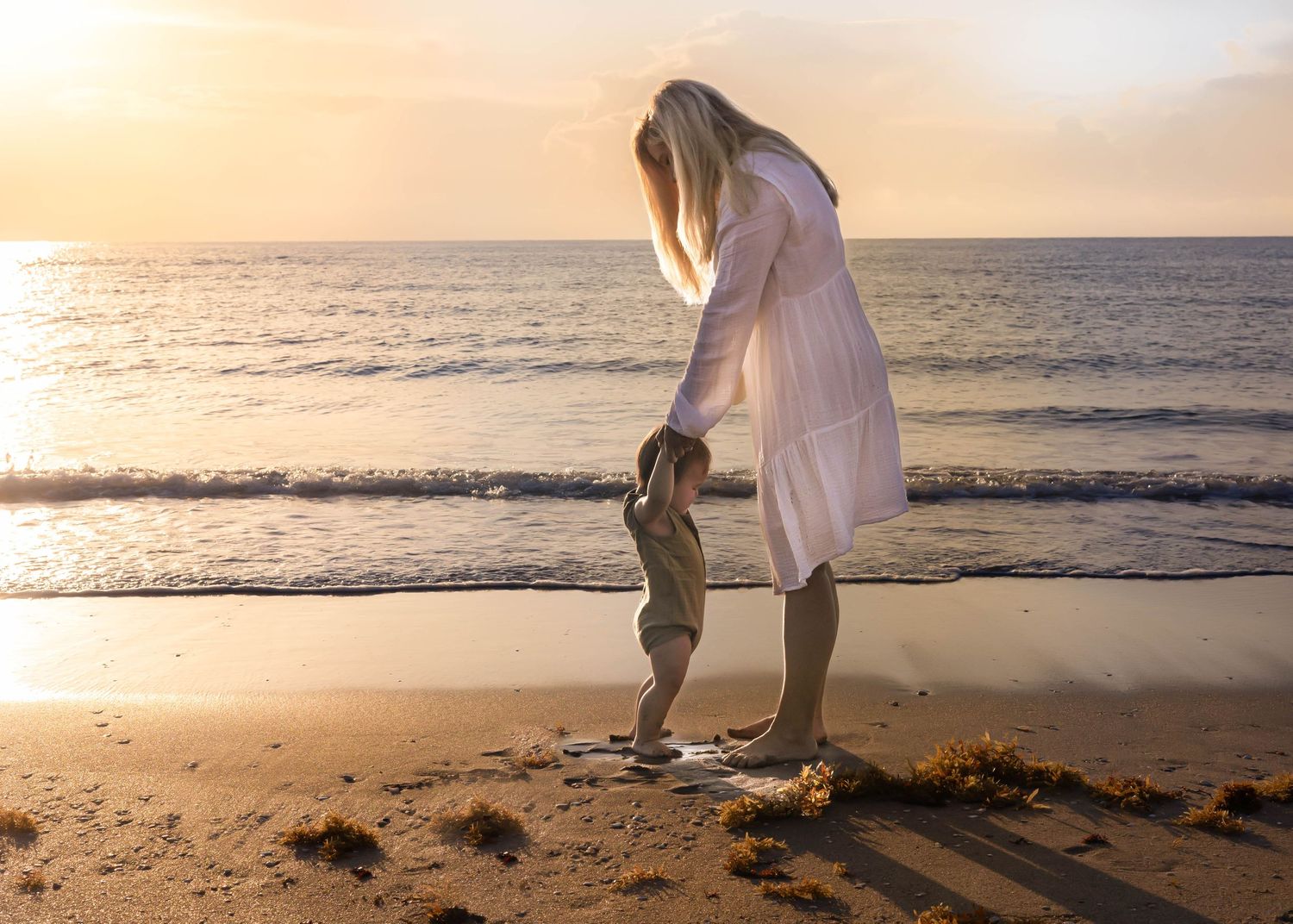 A mother holds her toddler's hands on a sandy beach at golden sunset, waves gently lapping the shore behind them.