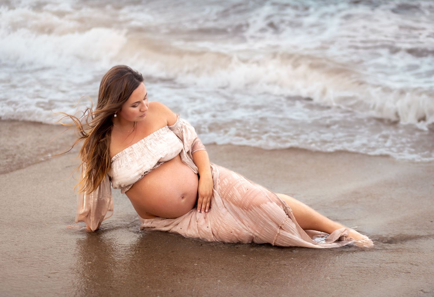 Pregnant woman in white lace top and pink skirt poses on sandy beach with ocean waves.