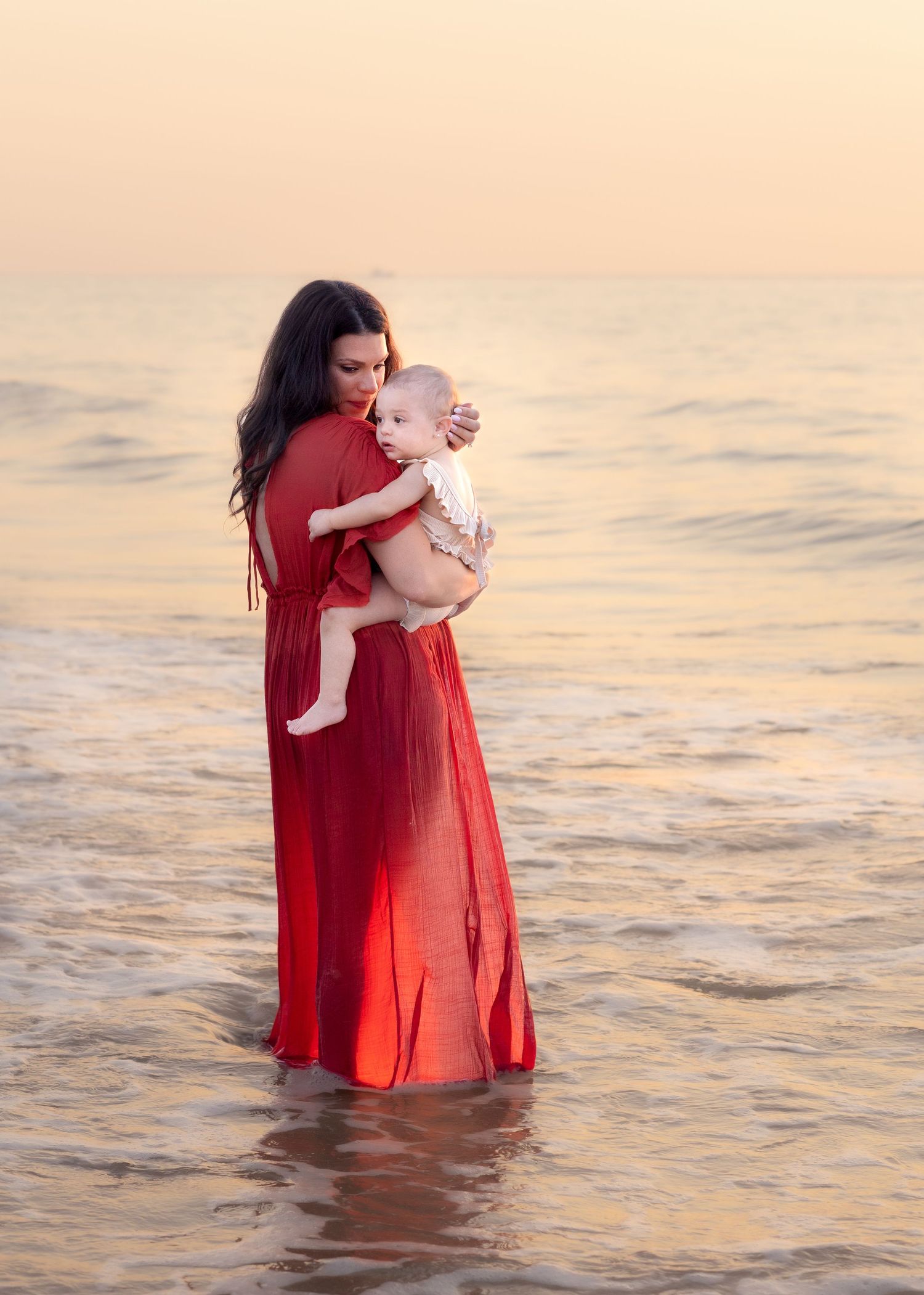 A mother in a red dress holds her baby while standing in shallow water at the golden bathed beach at sunrise.