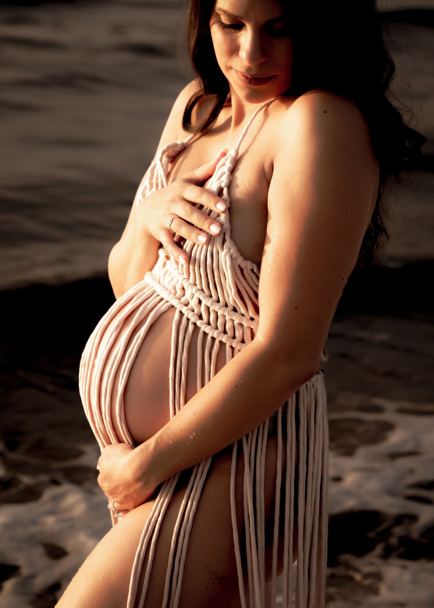 Pregnant woman in macrame dress cradling baby bump at beach during golden hour maternity photoshoot.