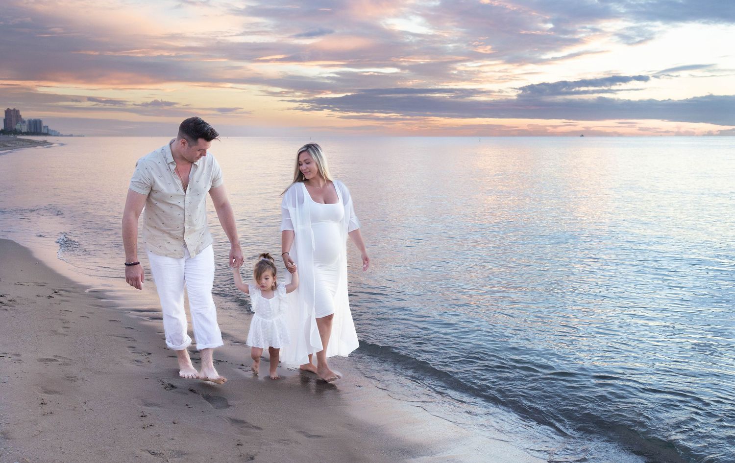 Family of three in white outfits walking on a beach at sunset, pregnant mother holding young child's hand.