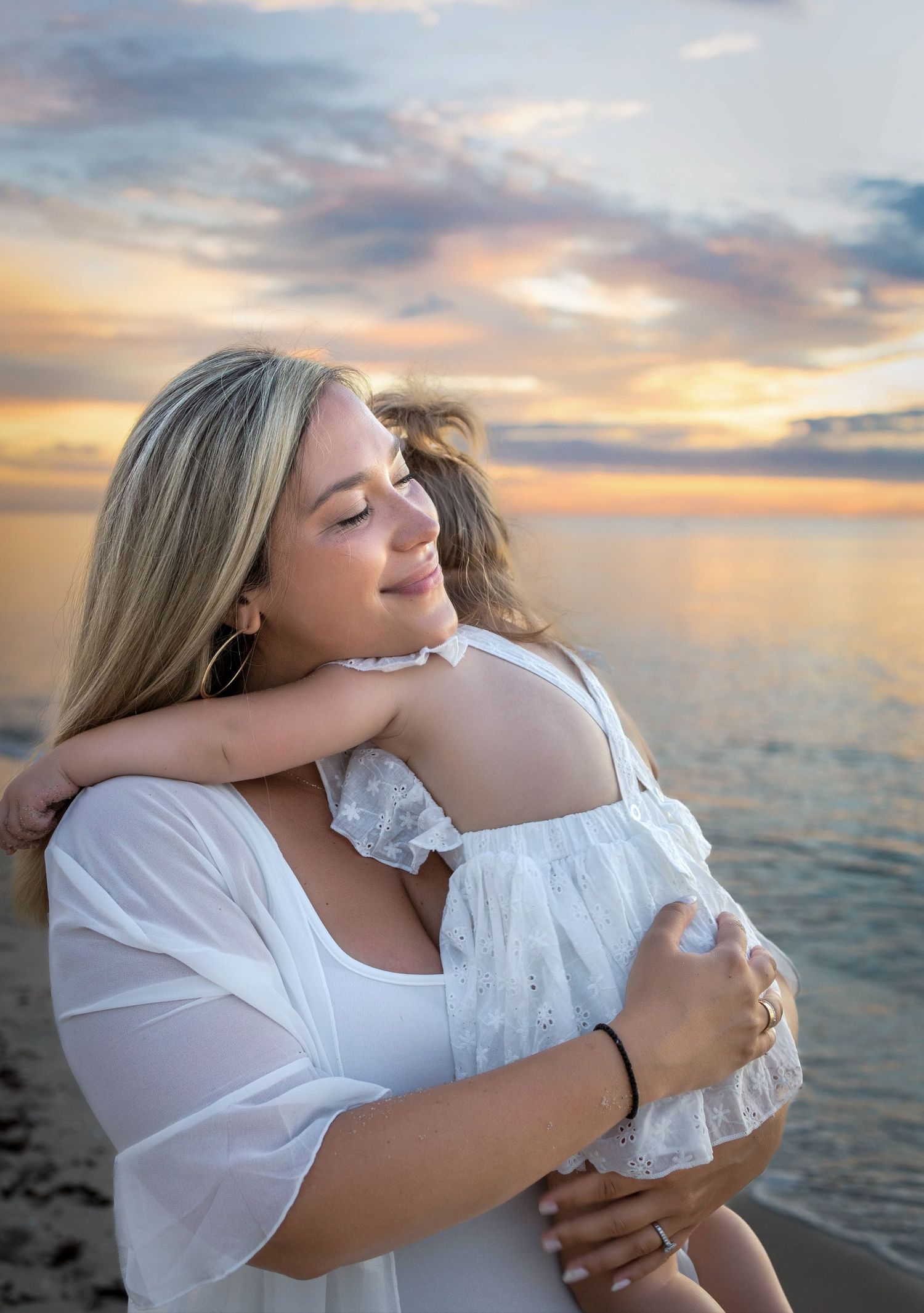 A mother tenderly hugs her child on a beach at sunset, both wearing white dresses with golden sky behind them.