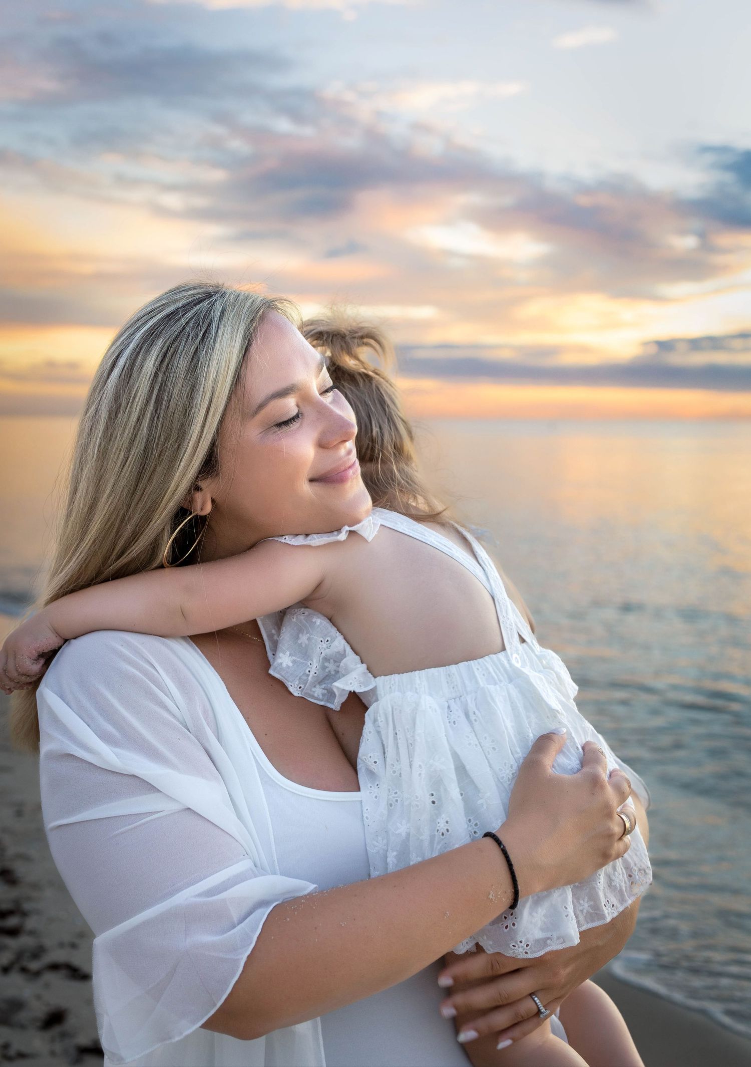 A mother in white dress hugs her child at a beach during a golden sunset over calm ocean waters.
