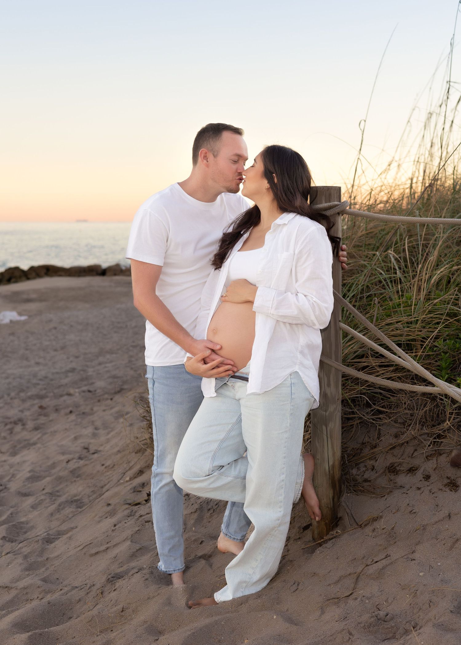 Couple sharing a kiss on sandy beach at sunset, man holding pregnant woman's belly near wooden fence.