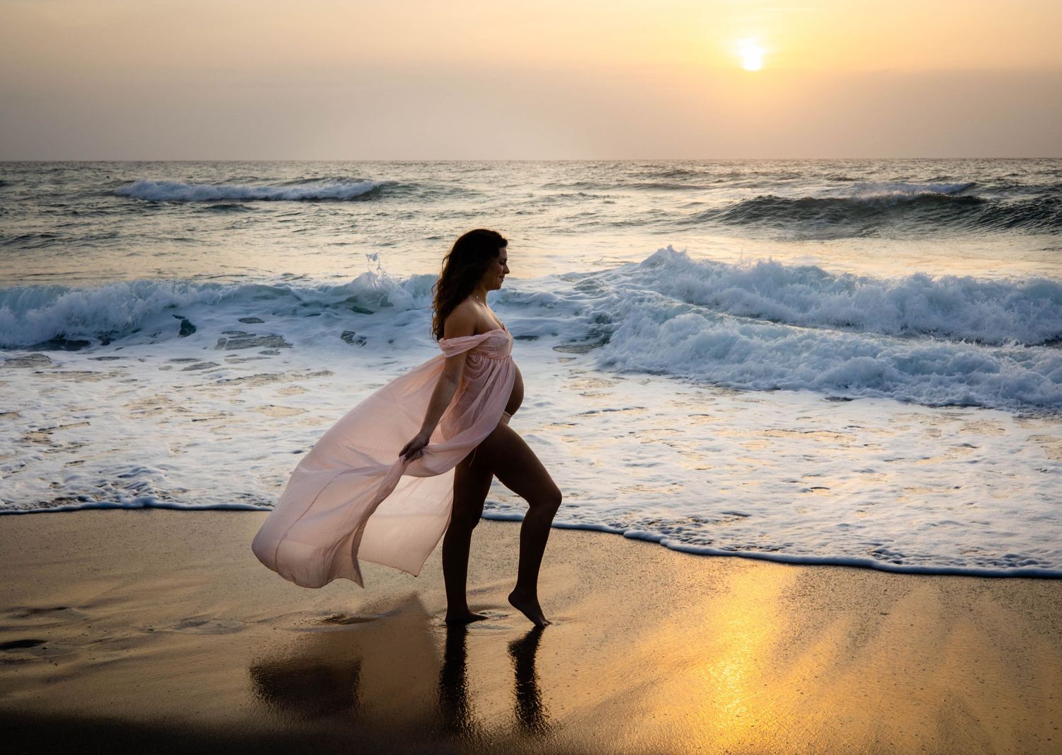 Pregnant woman in flowing pink dress standing on beach at sunset with ocean waves crashing behind her.