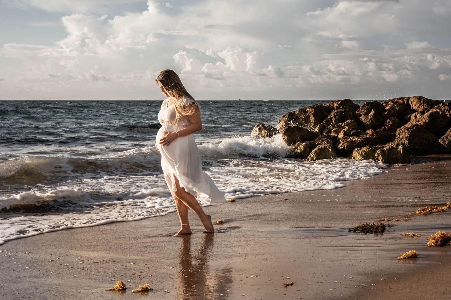 Pregnant woman in white flowing dress walking along the shoreline in Deerfield Beach.