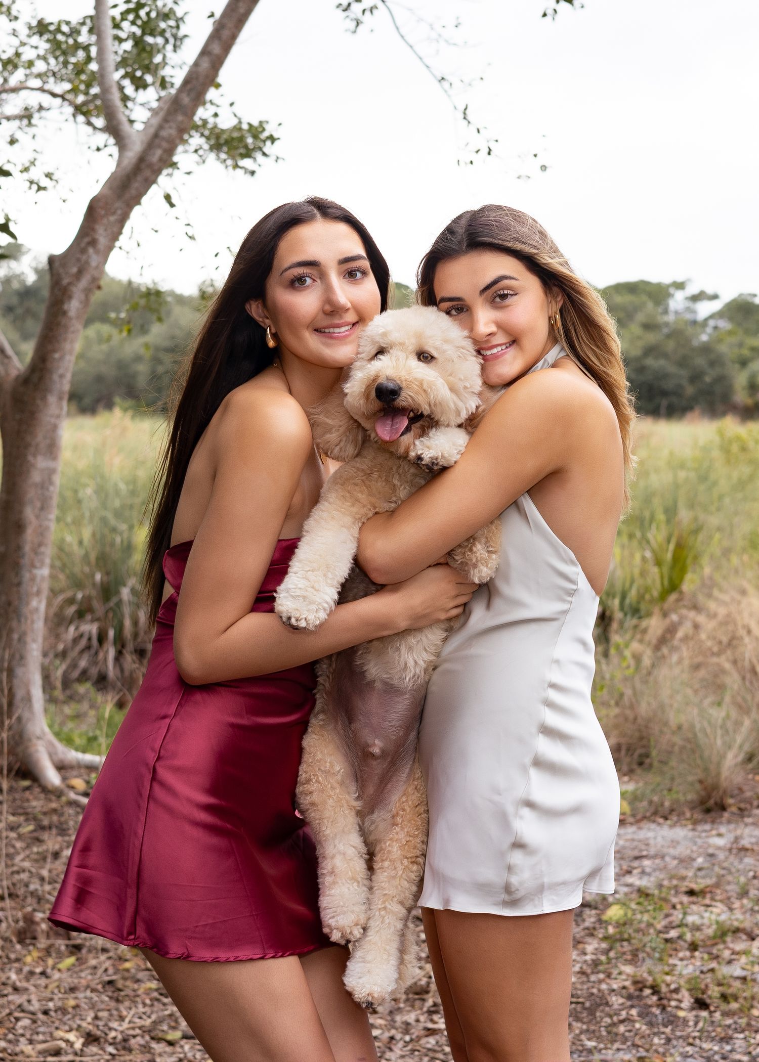 Two young women in elegant dresses smile while holding a fluffy golden doodle outdoors.