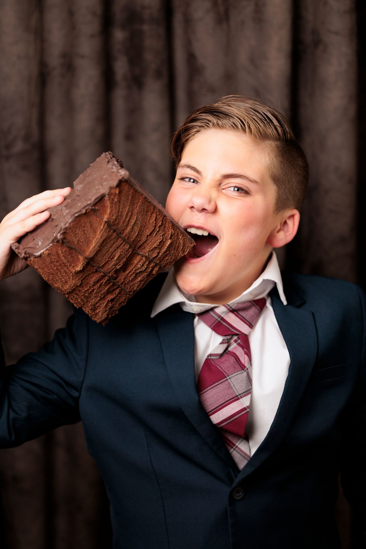 A boy in a navy suit pretends to bite an oversized chocolate bar prop in a theatrical production.