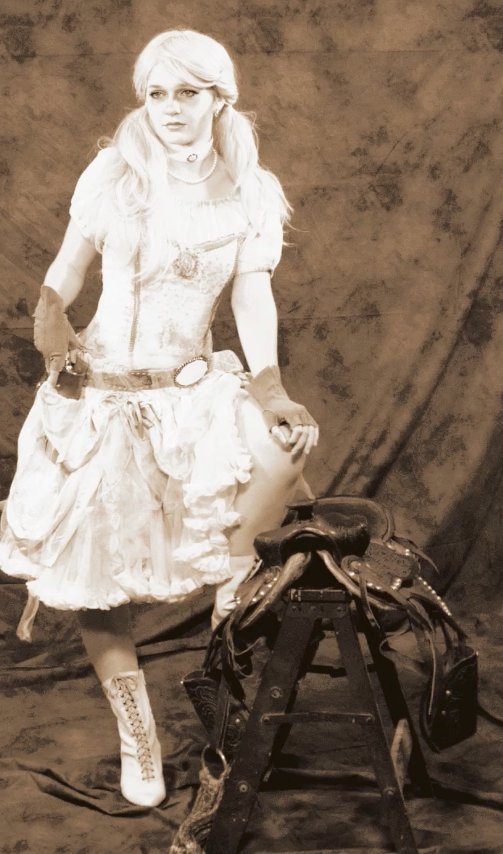 A woman in a white ruffled dress and boots poses beside a saddle in a sepia portrait.
