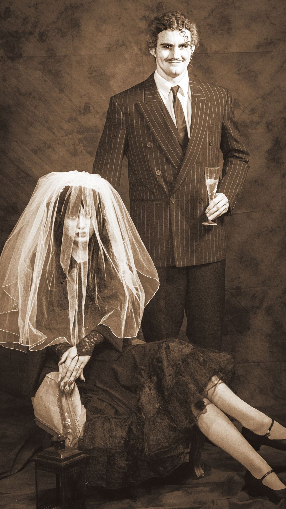 A sepia portrait of a bride sitting on the floor and a suited groom holding a champagne glass.