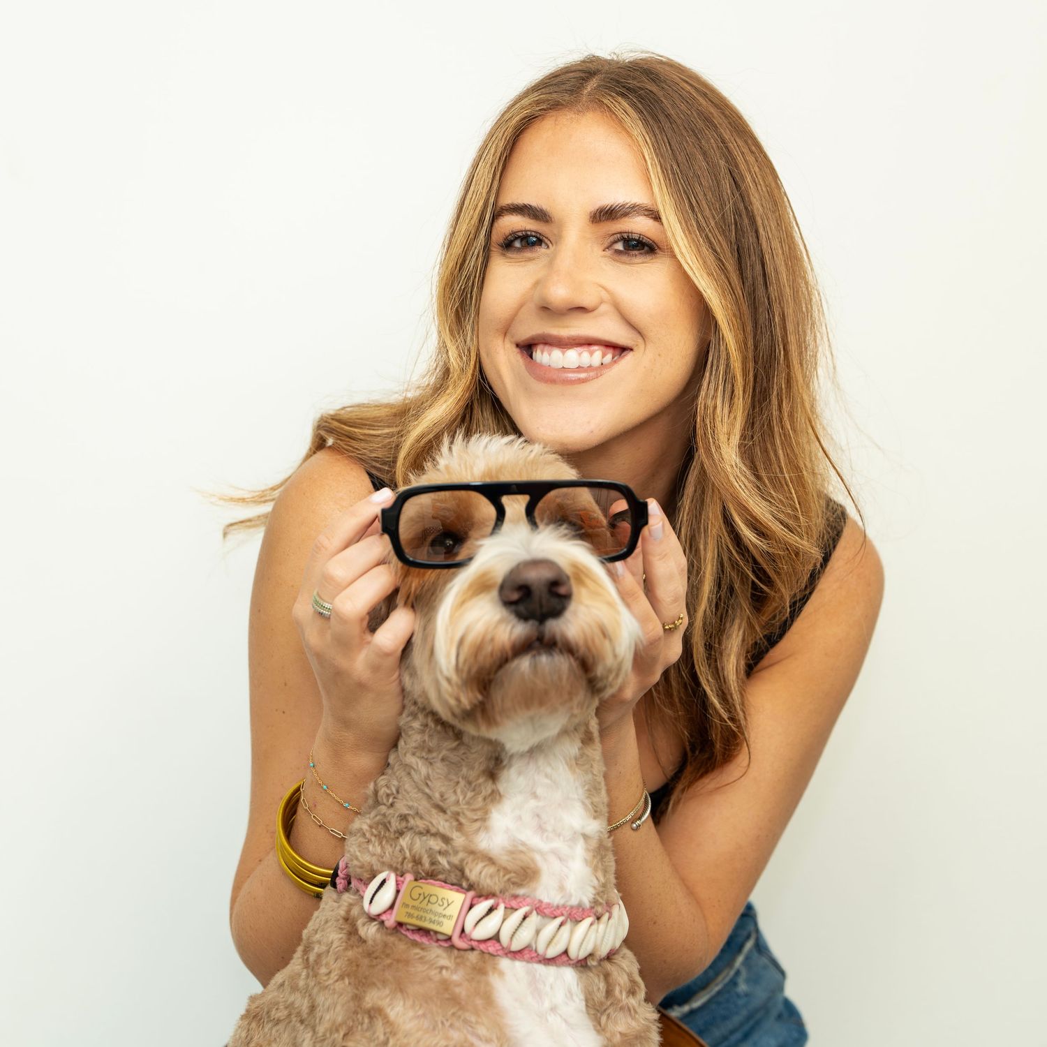 Smiling woman holding sunglasses on a fluffy dog wearing a shell collar against a white background.