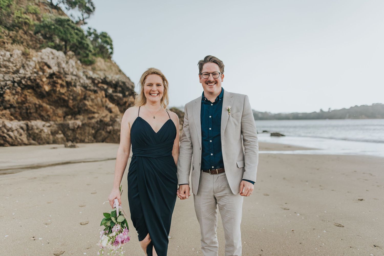 A couple in white and pink attire poses joyfully in a green outdoor setting on a cloudy day.