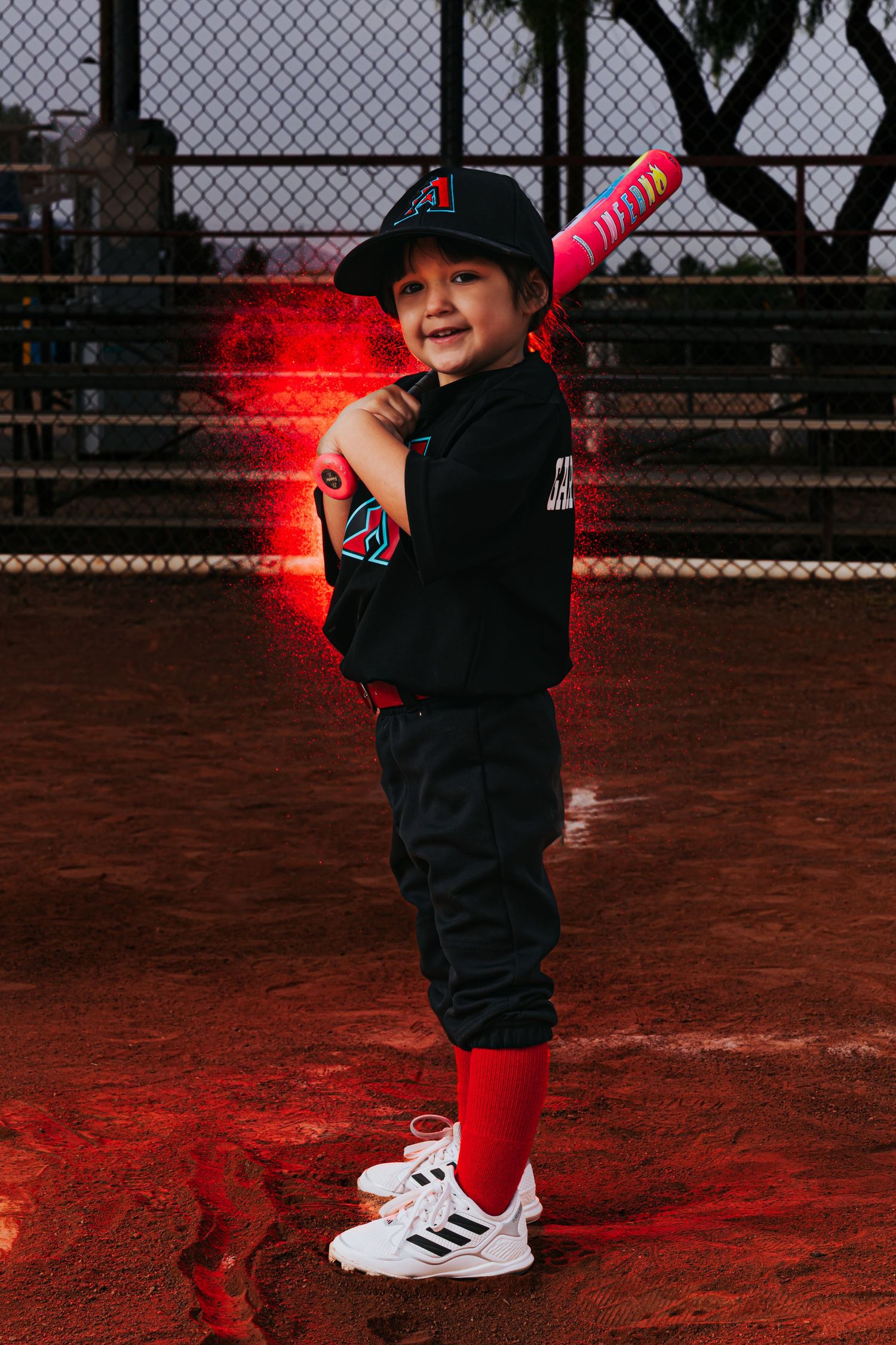 Young baseball player in black uniform holds pink bat on red-lit dirt field, smiling confidently in white Adidas cleats.