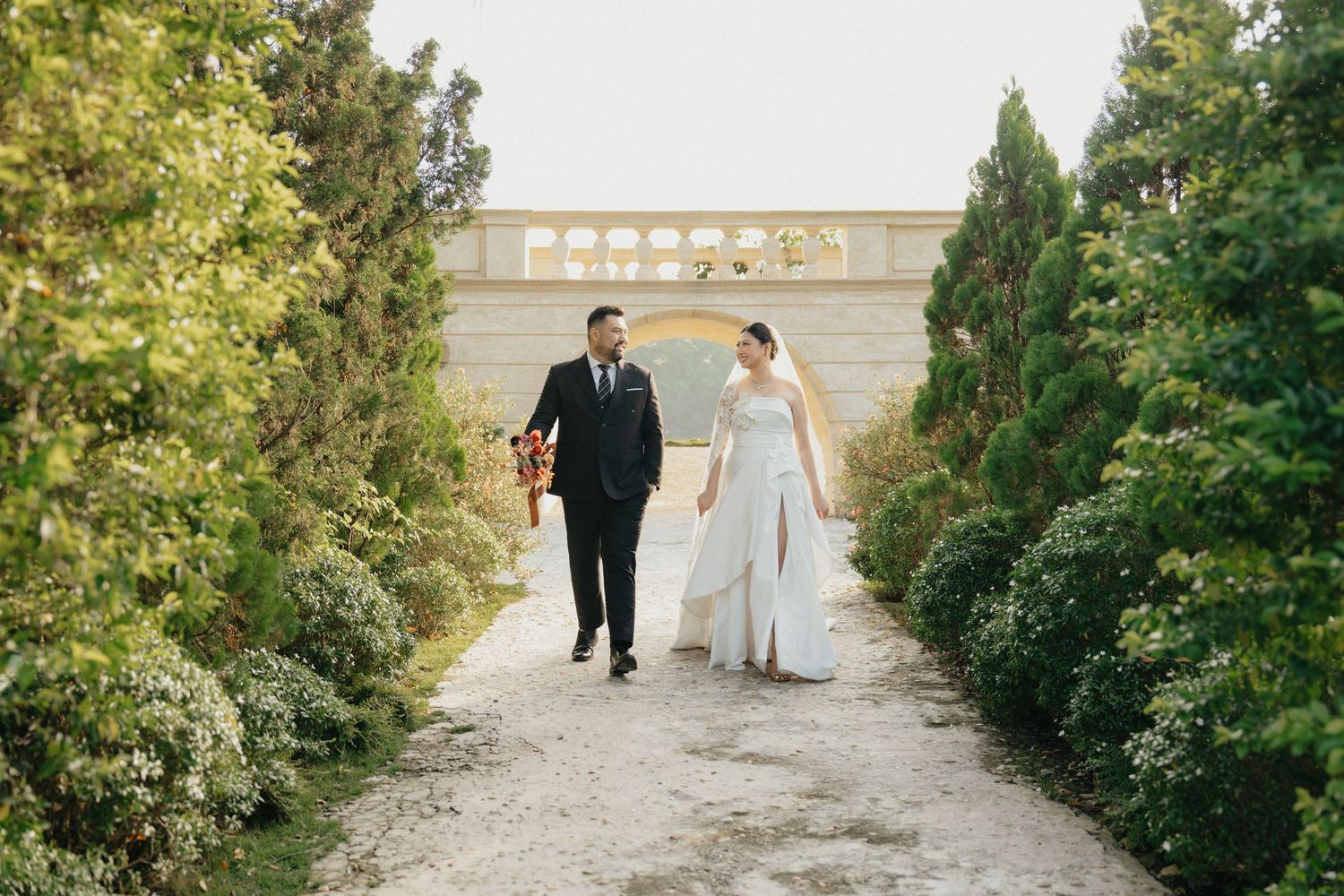 Bride and groom walking hand in hand along a garden path lined with tall green trees.