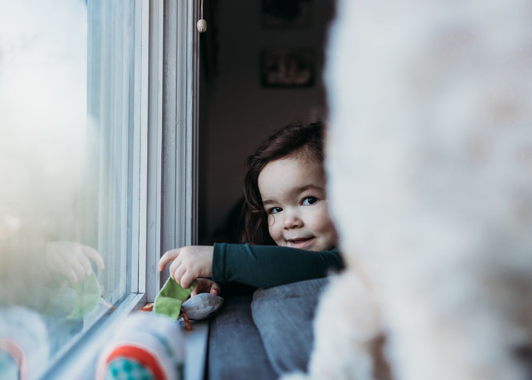 A small child peeks playfully from behind a window while holding a toy.