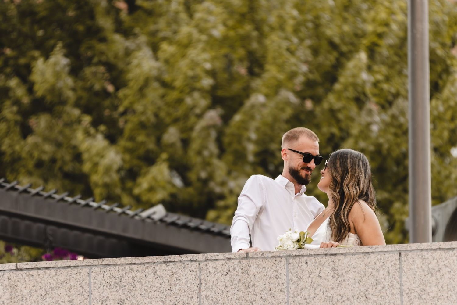 A romantic couple shares an intimate moment on a concrete balcony with lush greenery in the background.