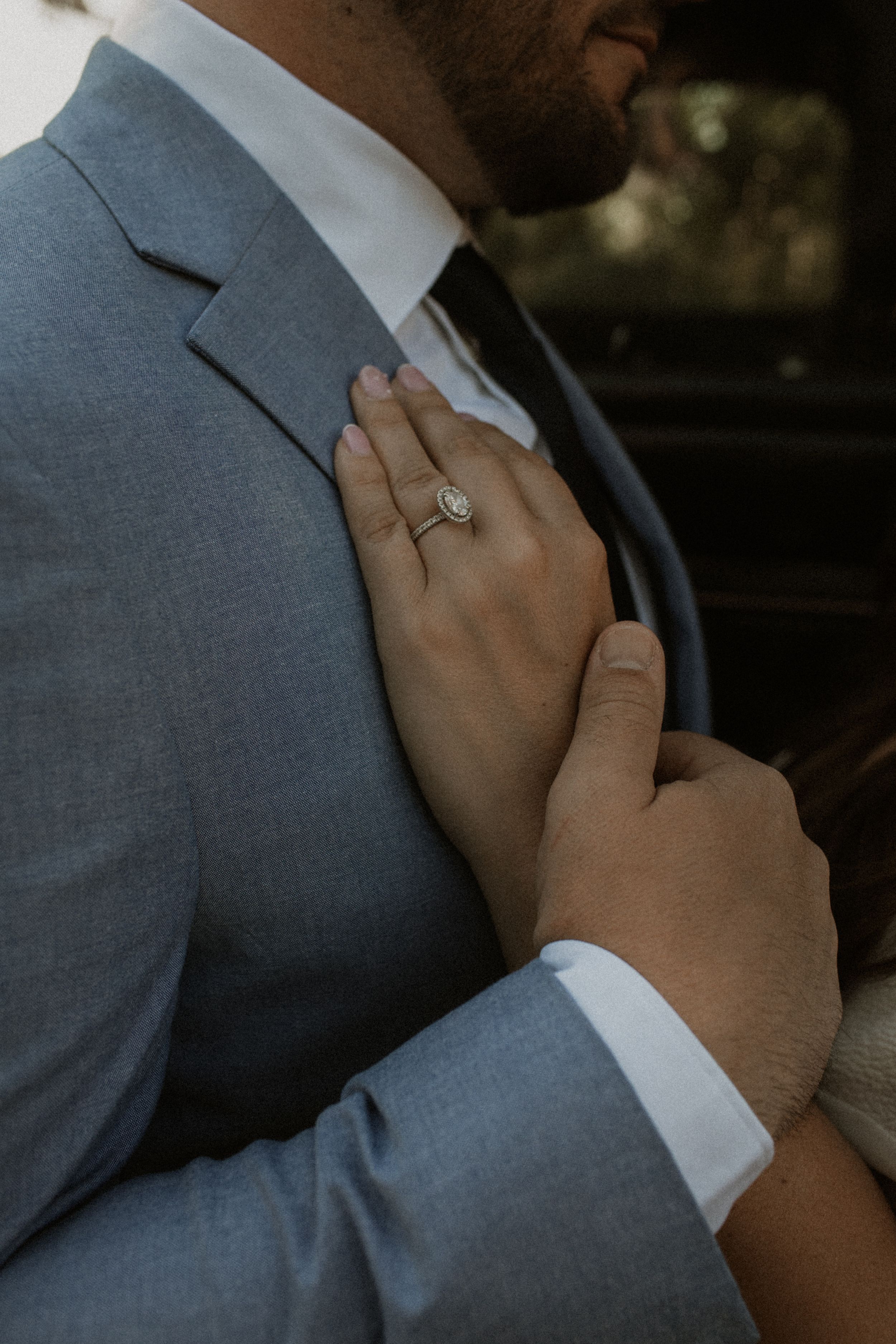 A hand wearing an engagement ring gently adjusts the collar of a grey suit and white dress shirt.