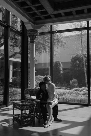 Black and white photo of couple sitting in sunlit porch with large windows and natural scenery outside.