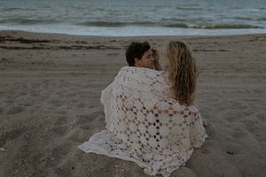 A couple shares an intimate moment on a sandy beach at sunset with the ocean waves in the background.