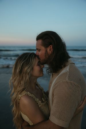 A romantic couple embraces on the beach at sunset with ocean waves in the background.