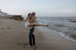 A couple shares a romantic moment walking along a sandy beach at sunset with waves crashing in the background.
