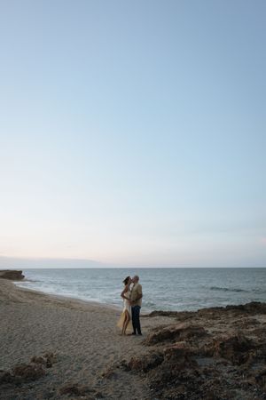 Two figures stand together on a rocky beach at dusk with the ocean and calm sky in the background.