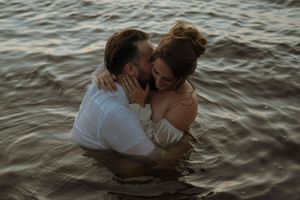 A romantic couple embraces while standing in ocean water at sunset, creating a silhouetted scene against rippling waves.