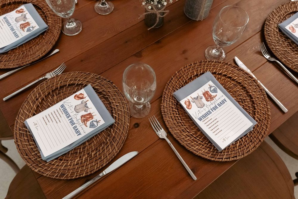 Baby shower table setting with woven chargers, crystal glasses, and Wishes for Baby cards on wooden table.