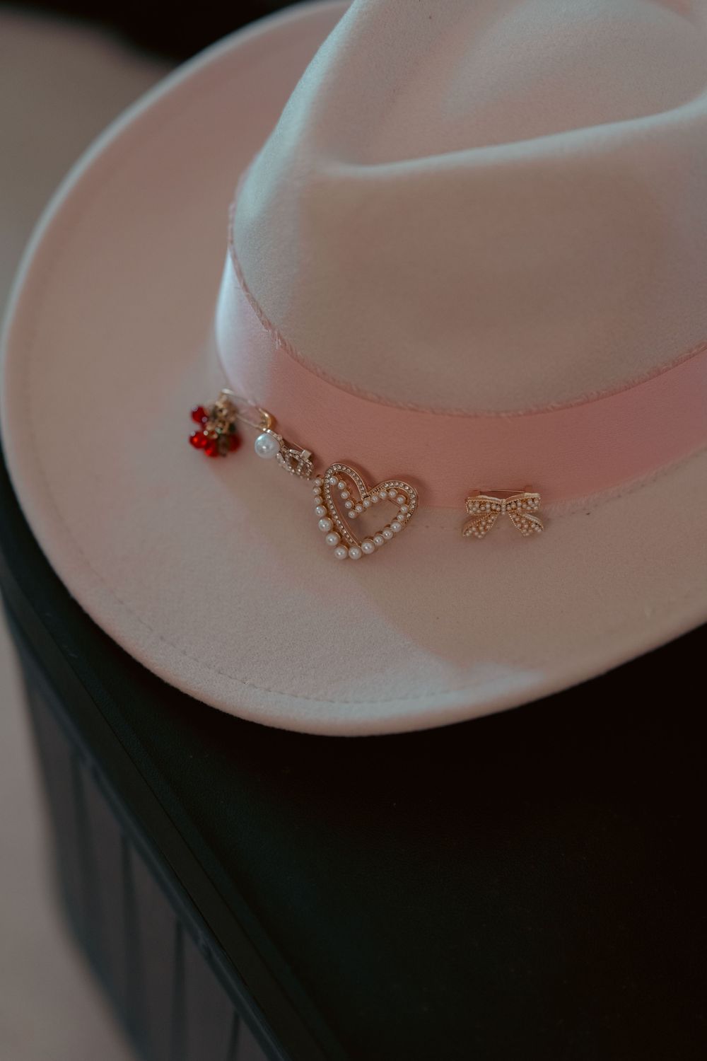 Pink felt fedora hat adorned with pearl heart, bow, and floral charm pins.