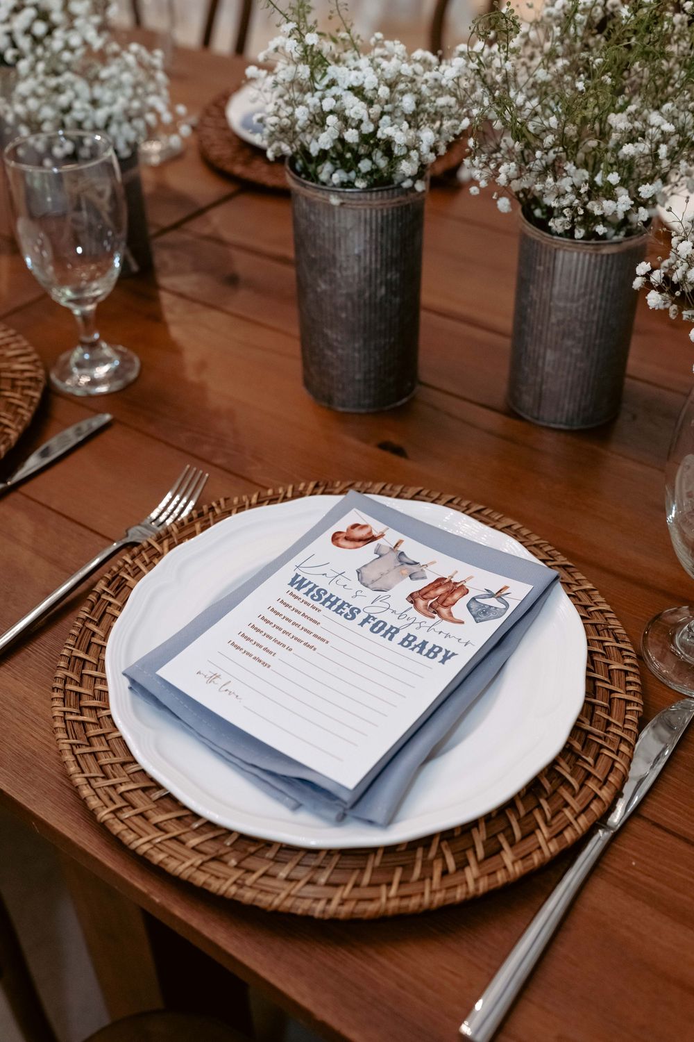 Close-up of baby shower place setting with Wishes for Baby card, white plate, woven charger, and floral centerpiece.