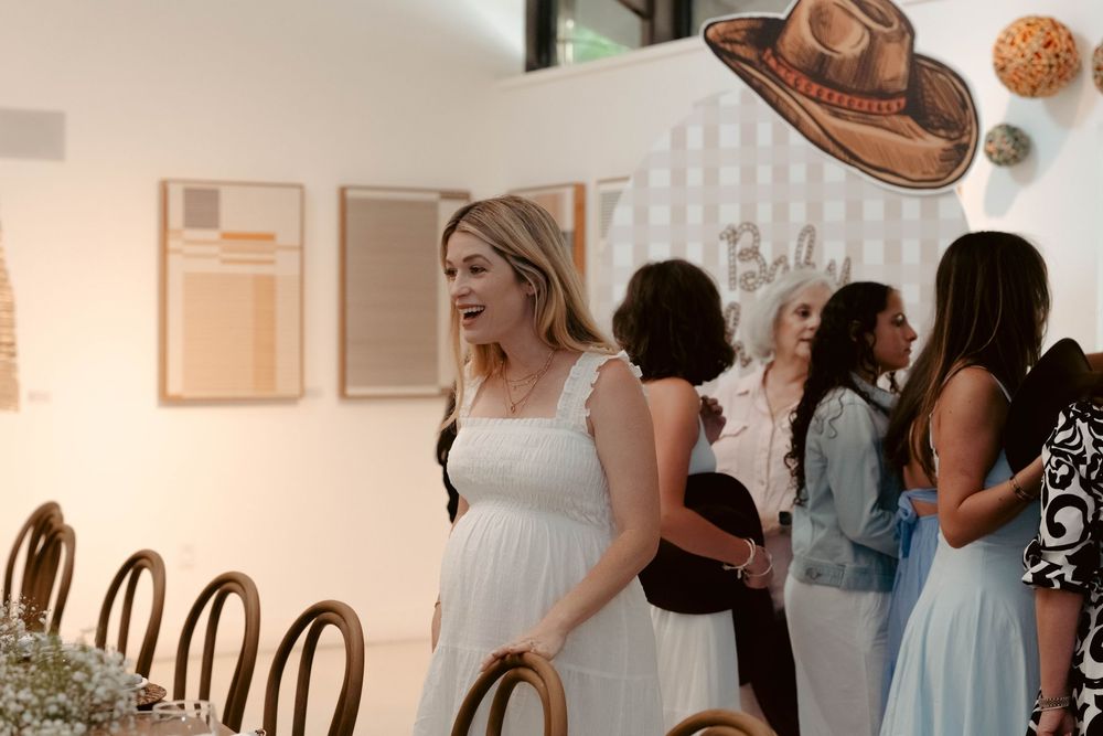 Pregnant woman in white dress mingles with guests at western-themed Baby Schroeder baby shower event.