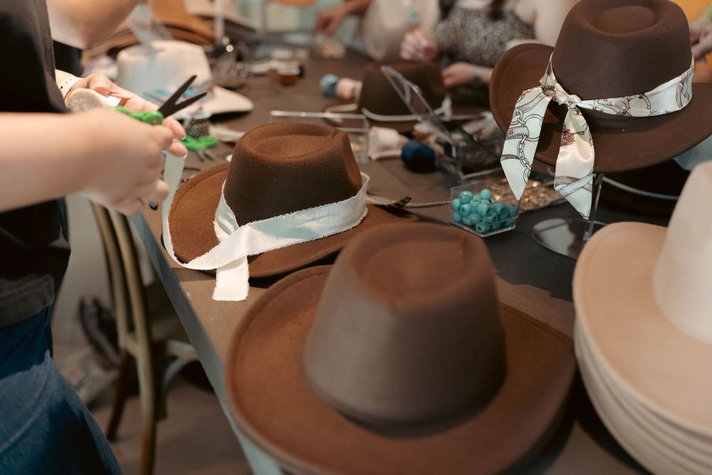 Hands decorating brown felt cowboy hats with ribbon and accessories at a craft table.