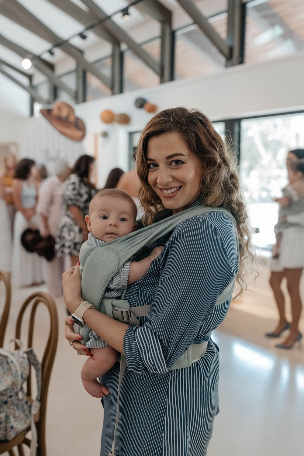 Smiling woman in striped dress holding infant at an indoor baby shower event.