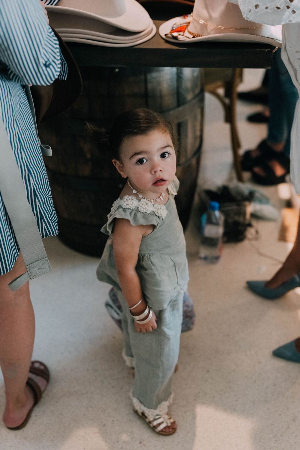 Adorable toddler girl in sage green jumpsuit standing near a wooden barrel at a party.