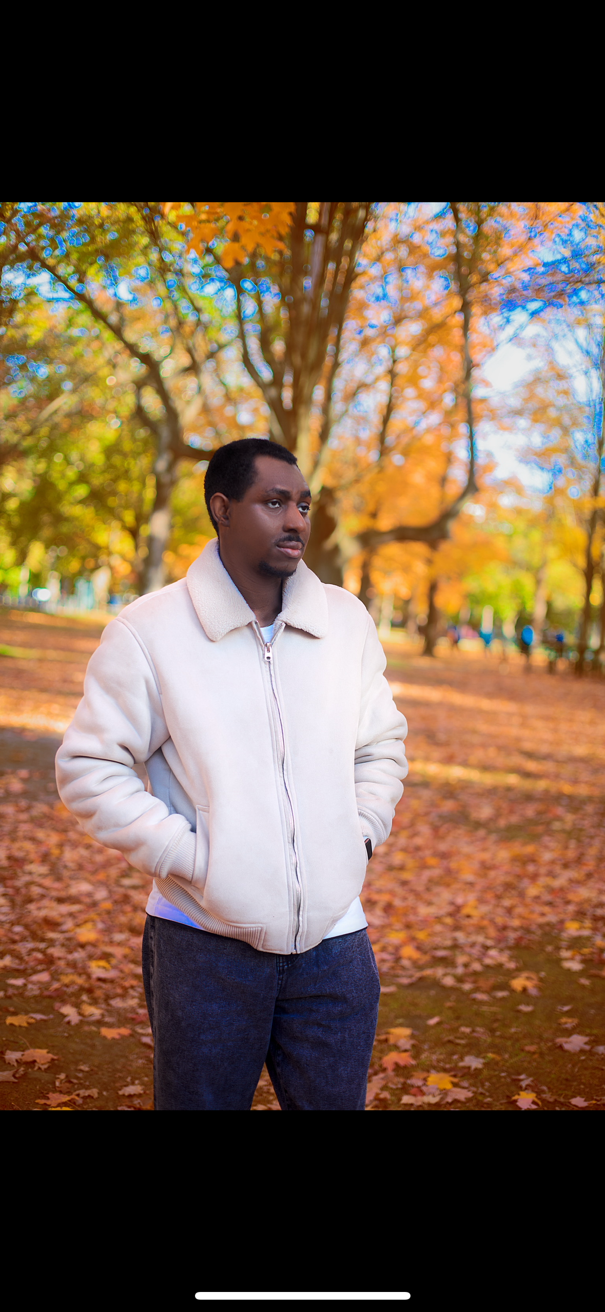 Black man in cream sherpa jacket standing in autumn park with colorful orange and yellow foliage.