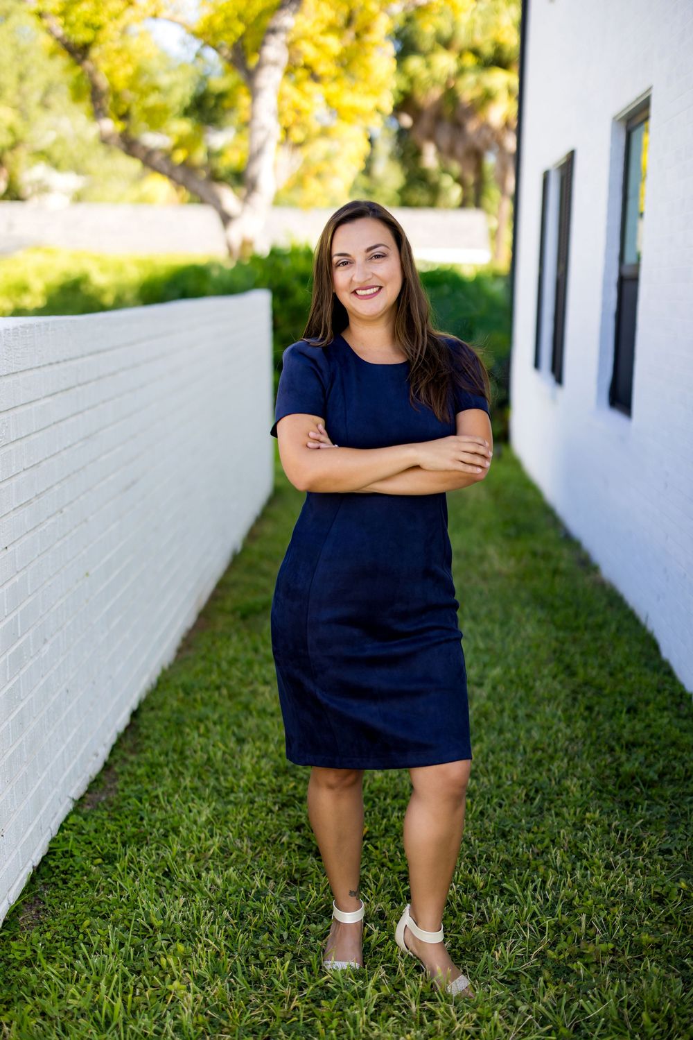 Woman in navy dress stands confidently with arms crossed on green lawn between white walls.
