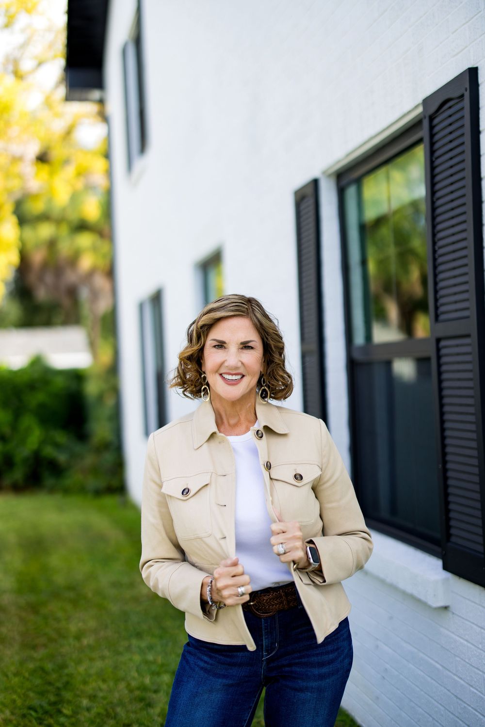 Smiling woman in beige jacket and jeans standing outdoors beside a white brick building with black shutters.