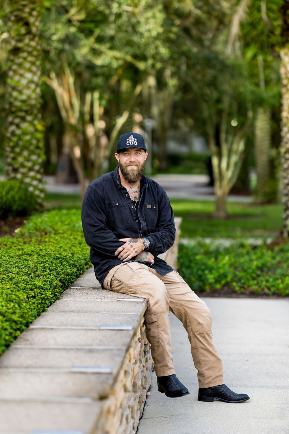 Bearded man in black cap and jacket sits on stone wall in lush green park setting.