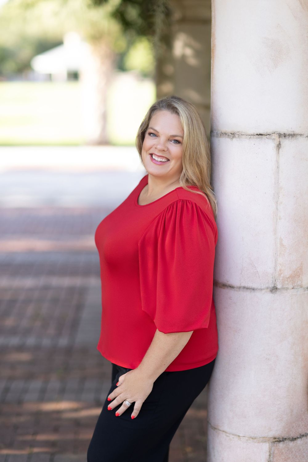 Smiling blonde woman in red top leaning against stone column outdoors.