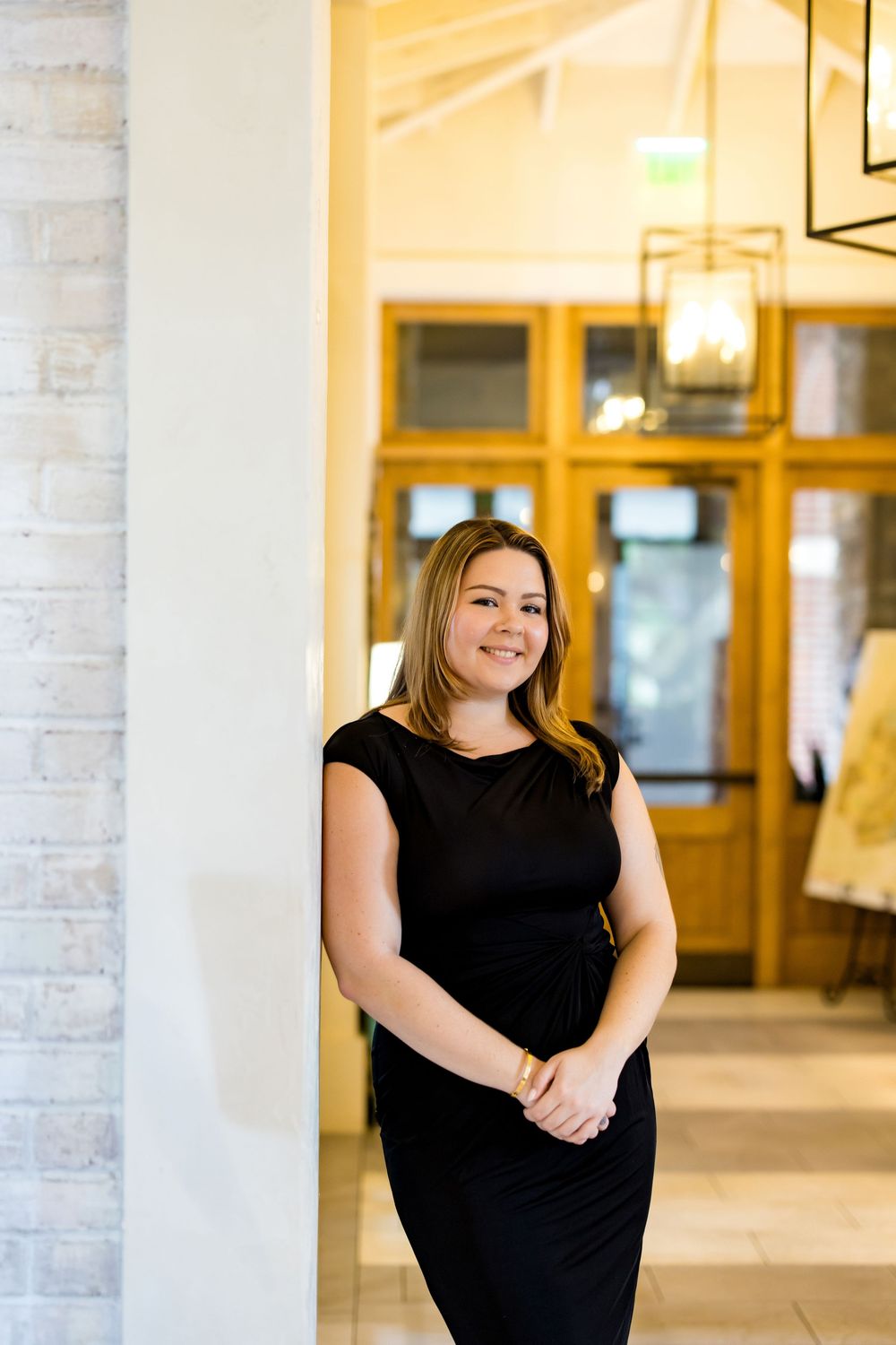 A smiling woman in a black dress leans against a white pillar in an elegant interior with warm lighting.