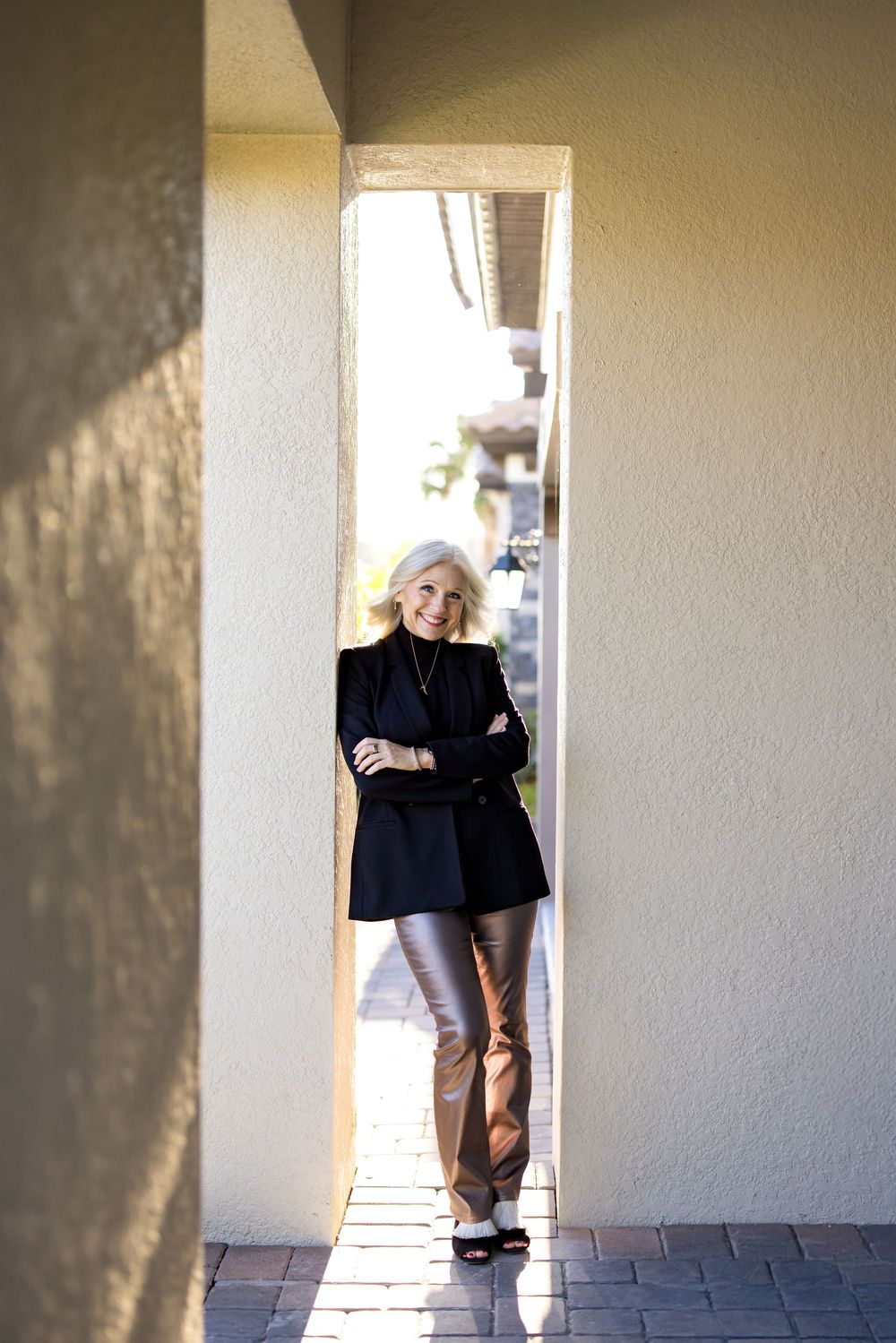 Elegant blonde woman in black blazer and gold pants leaning against white stucco wall in sunny courtyard.