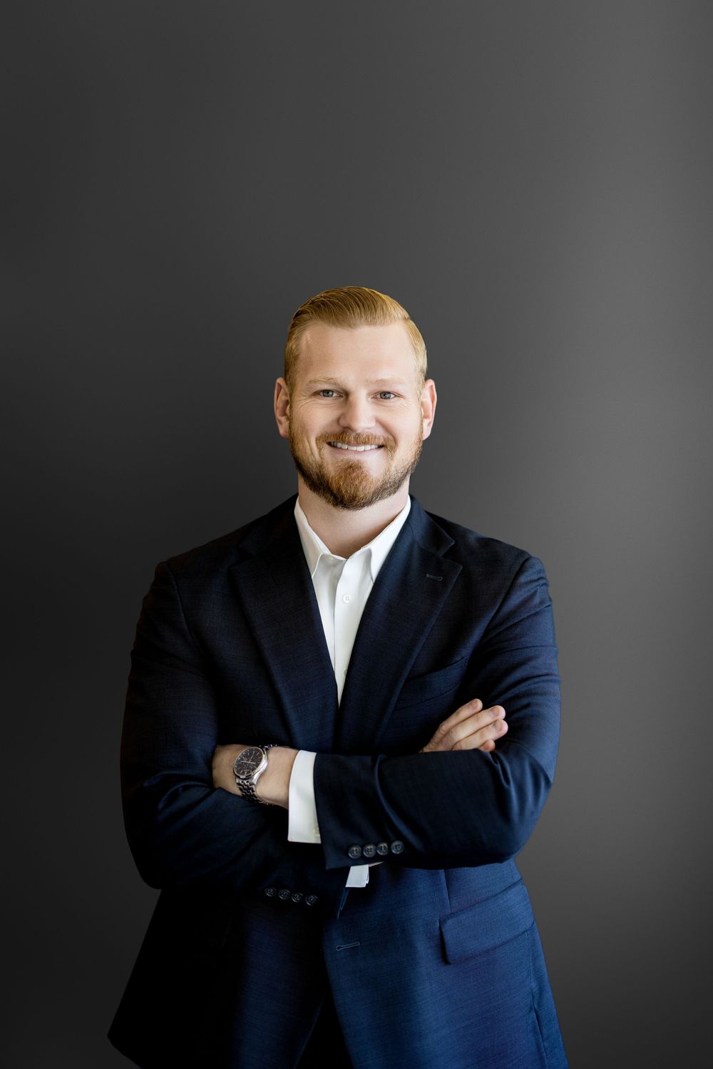Smiling redhead businessman in navy suit with arms crossed against dark gray background.