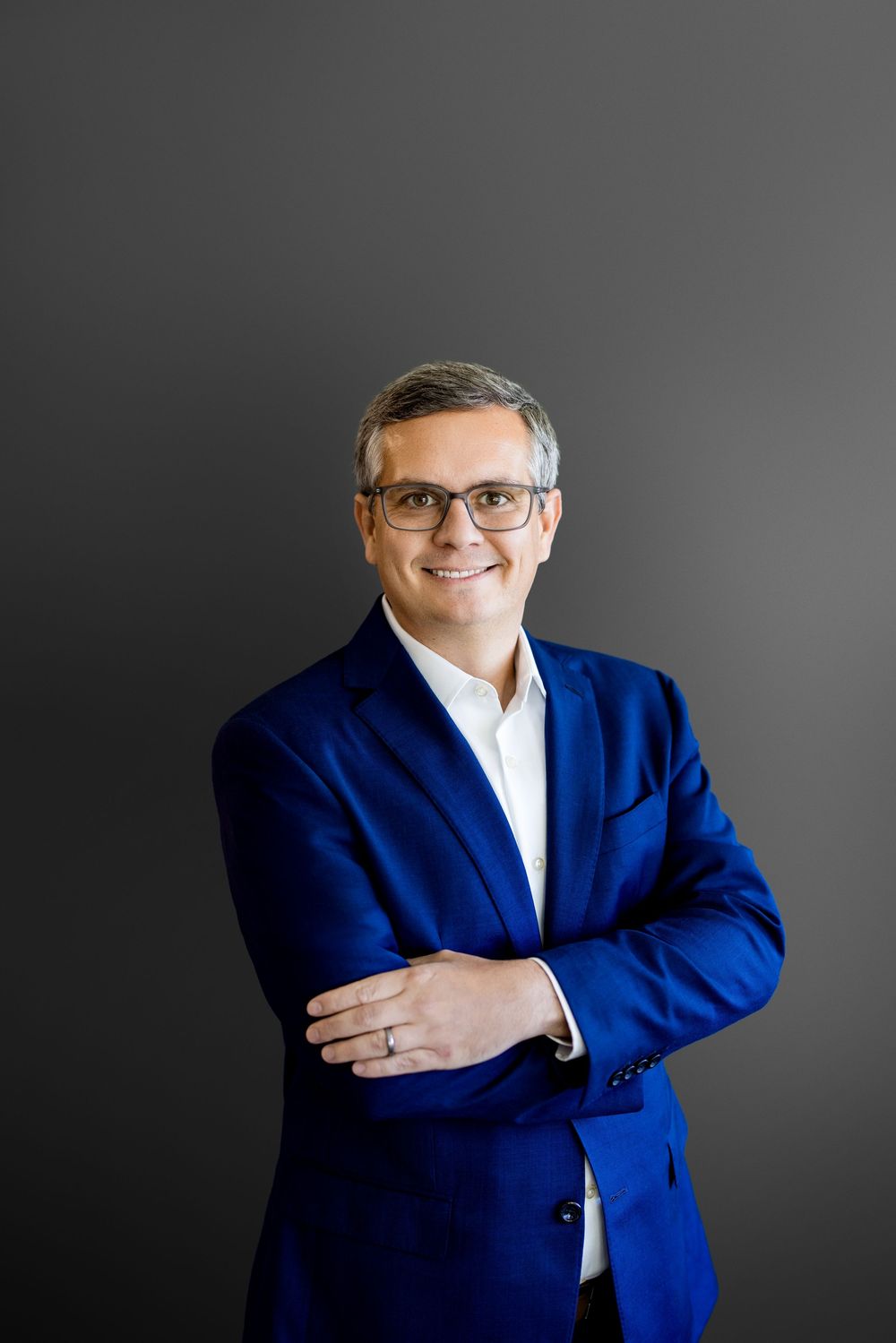 Professional man in blue blazer and glasses smiling with arms crossed against gray background.