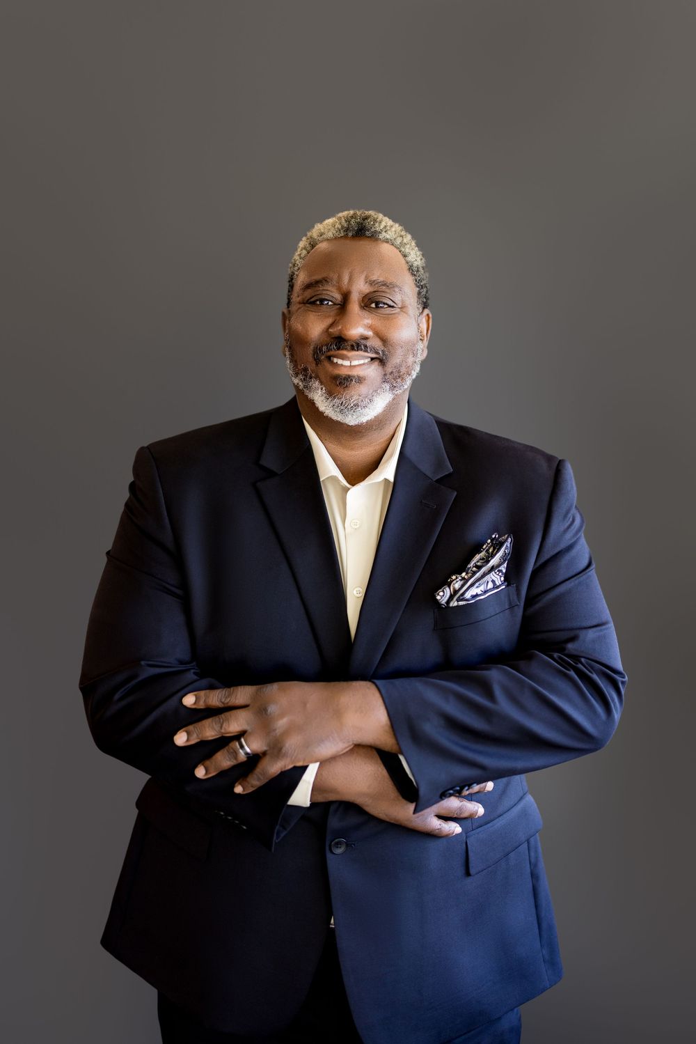 Smiling middle-aged Black man in navy blazer with pocket square, arms crossed, posing confidently against gray background.