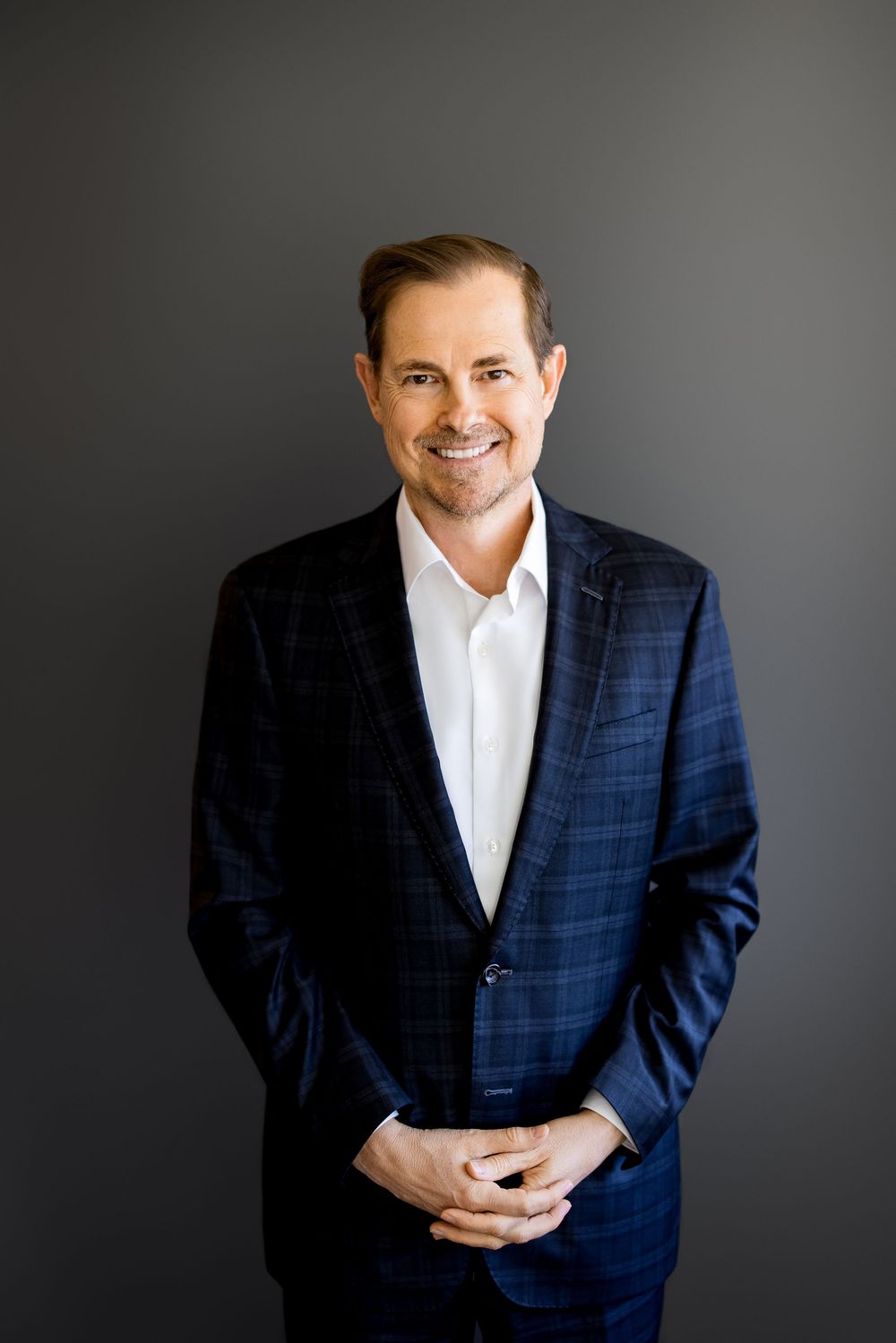 Professional man in navy plaid blazer smiling confidently against gray background in corporate headshot.