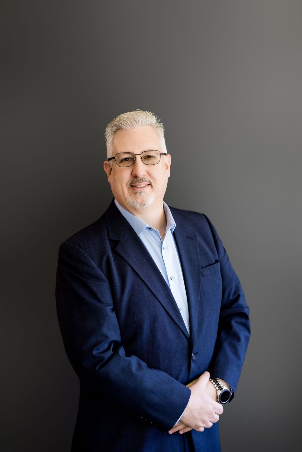 Professional man in navy blazer and glasses smiling confidently against a dark gray background.
