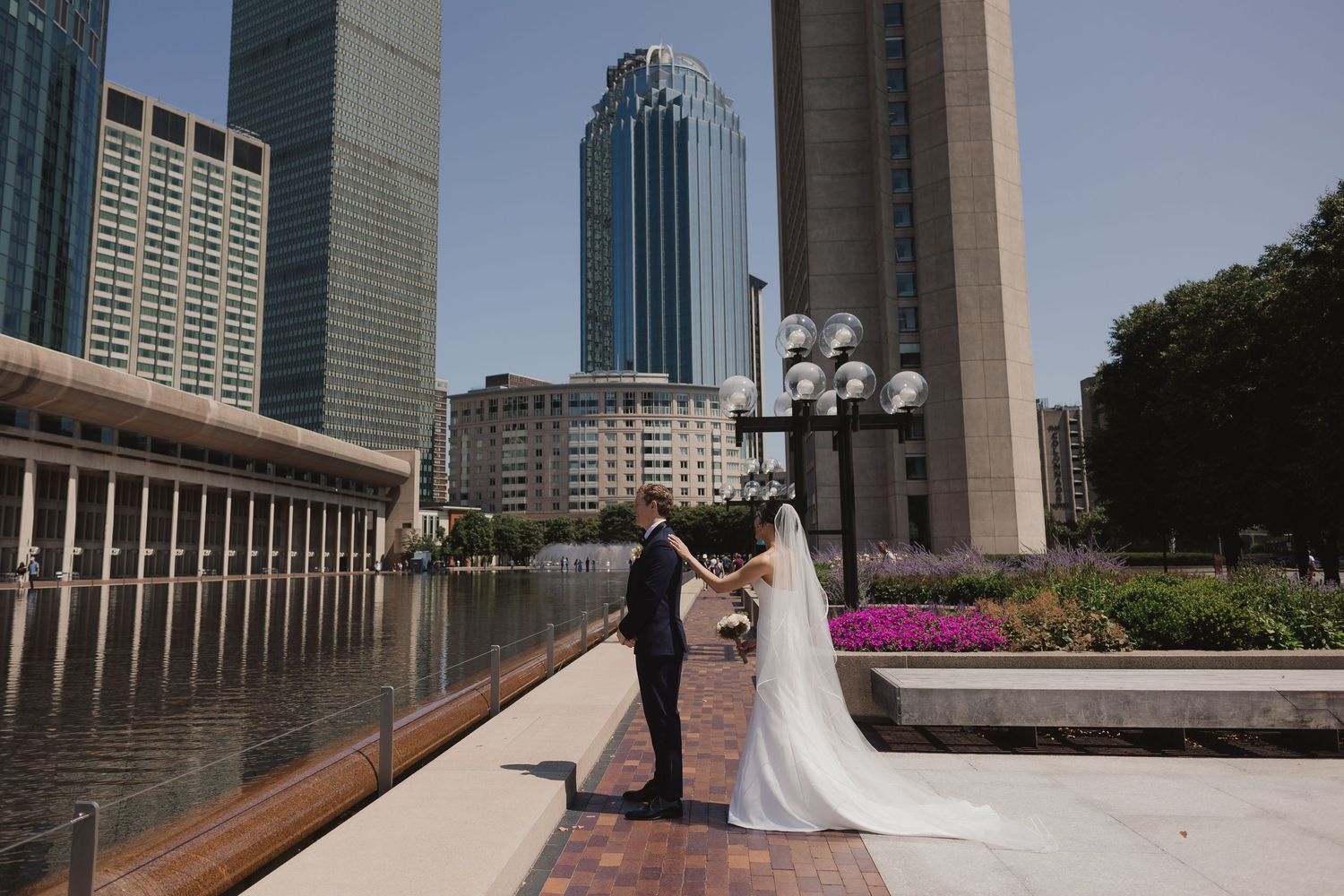 Bride and groom share a romantic moment on a waterfront promenade surrounded by city skyscrapers on their wedding day.